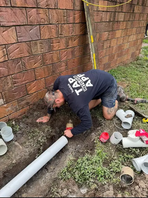 George kneeling in a trench beside a brick wall, with PVC drainage pipe and fittings laid out