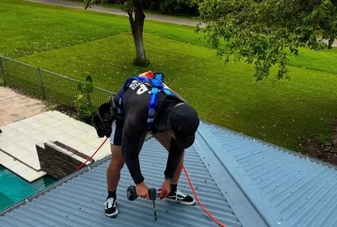A plumber in a safety harness drilling into a blue metal roof