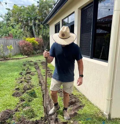 Mick in a straw hat with a shovel, standing in a freshly dug stormwater trench across a lawn