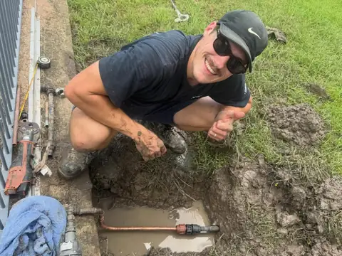 A smiling plumber crouched beside a muddy hole with a copper water pipe exposed